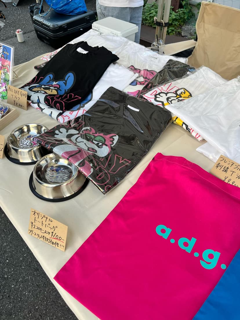 フリーマーケットのテーブルに並ぶ〈a.d.g.〉のTシャツ、トートバッグ、ステッカー